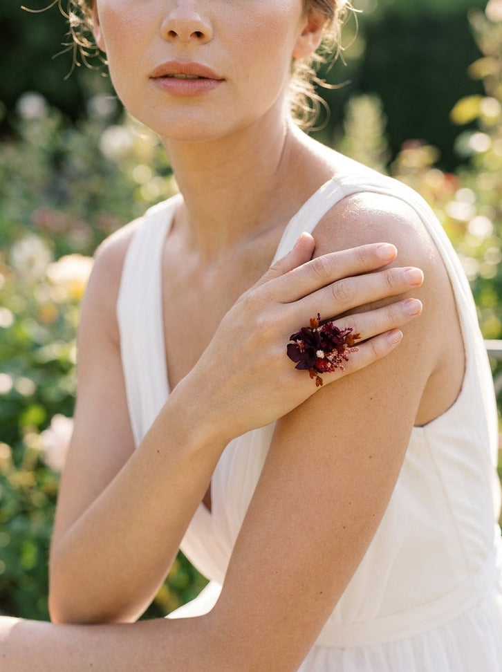 A woman in a white sleeveless dress wearing a dramatic statement ring with dark red flowers and gold-toned metal, posed outdoors against a soft green garden background.