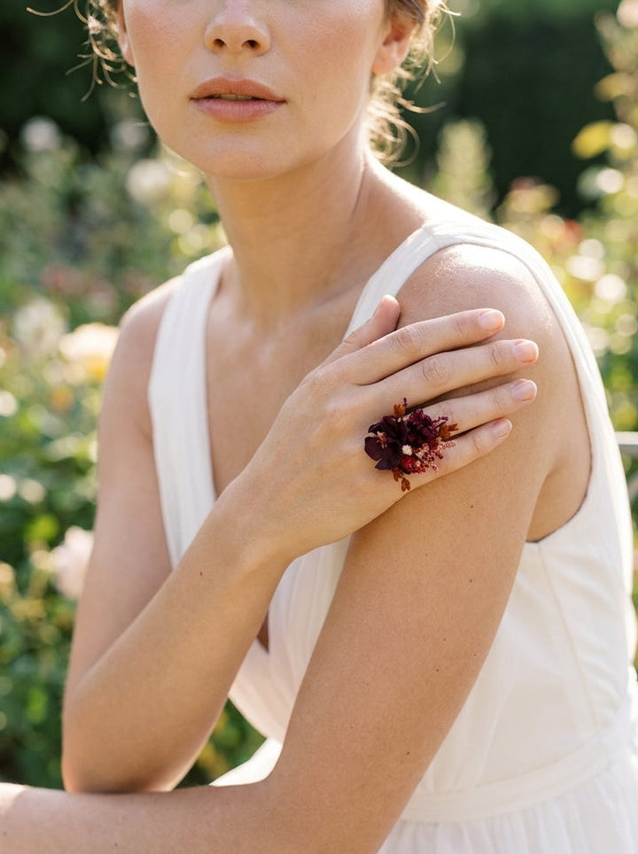 A woman in a white sleeveless dress wearing a dramatic statement ring with dark red flowers and gold-toned metal, posed outdoors against a soft green garden background.