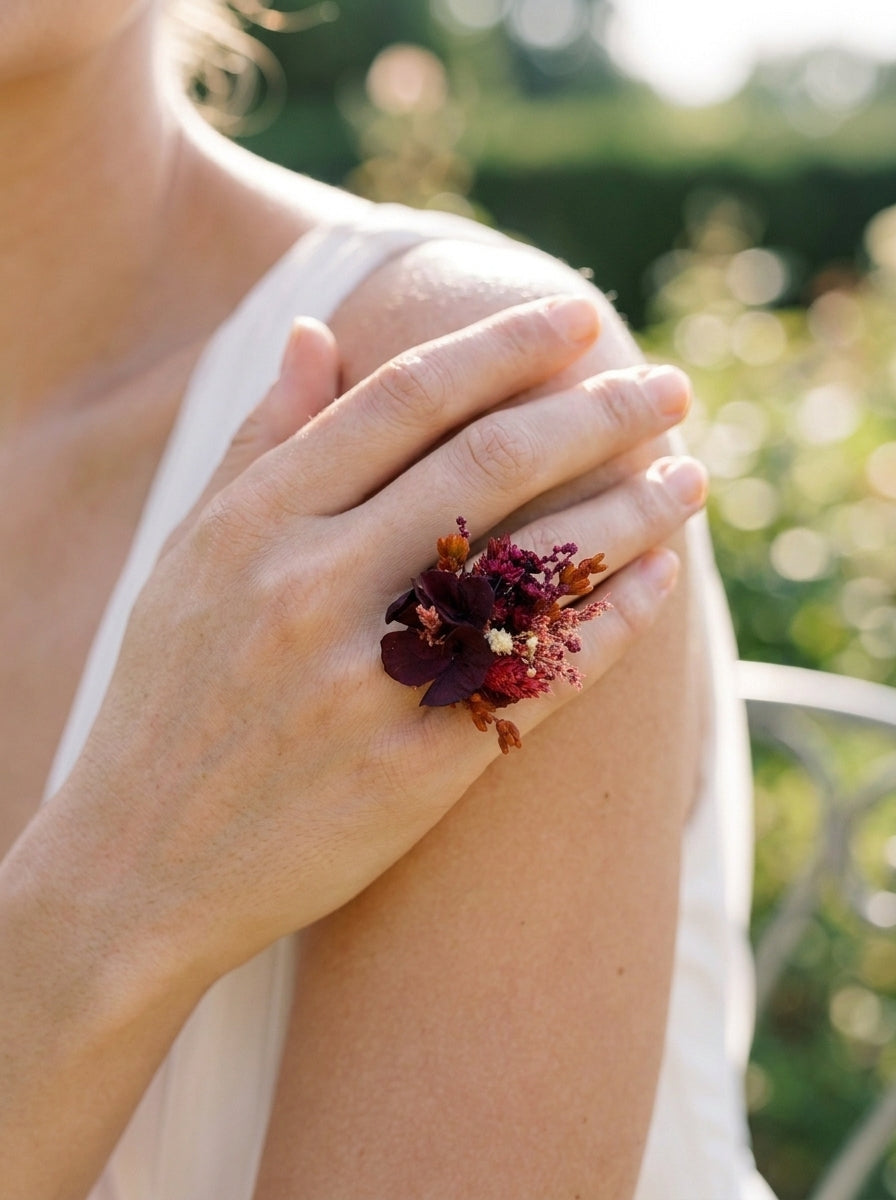 A close-up lifestyle shot of a woman wearing a small burgundy and dark red floral ring on her shoulder, with a soft-focus green garden background.