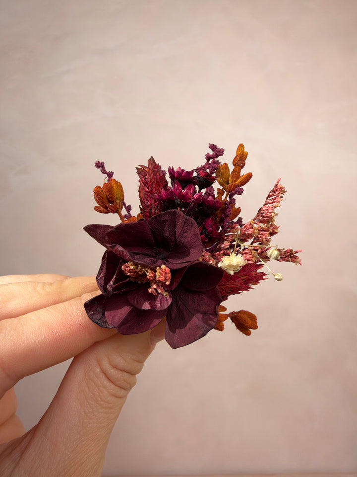 A hand holding a small dried floral arrangement featuring dark burgundy leaves, rust-orange seed pods, and tiny cream blossoms against a soft beige background.