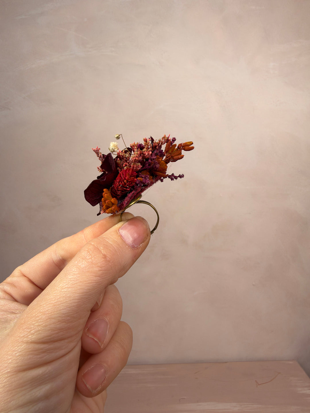 A hand holding a small dried flower ring against a soft beige textured background.