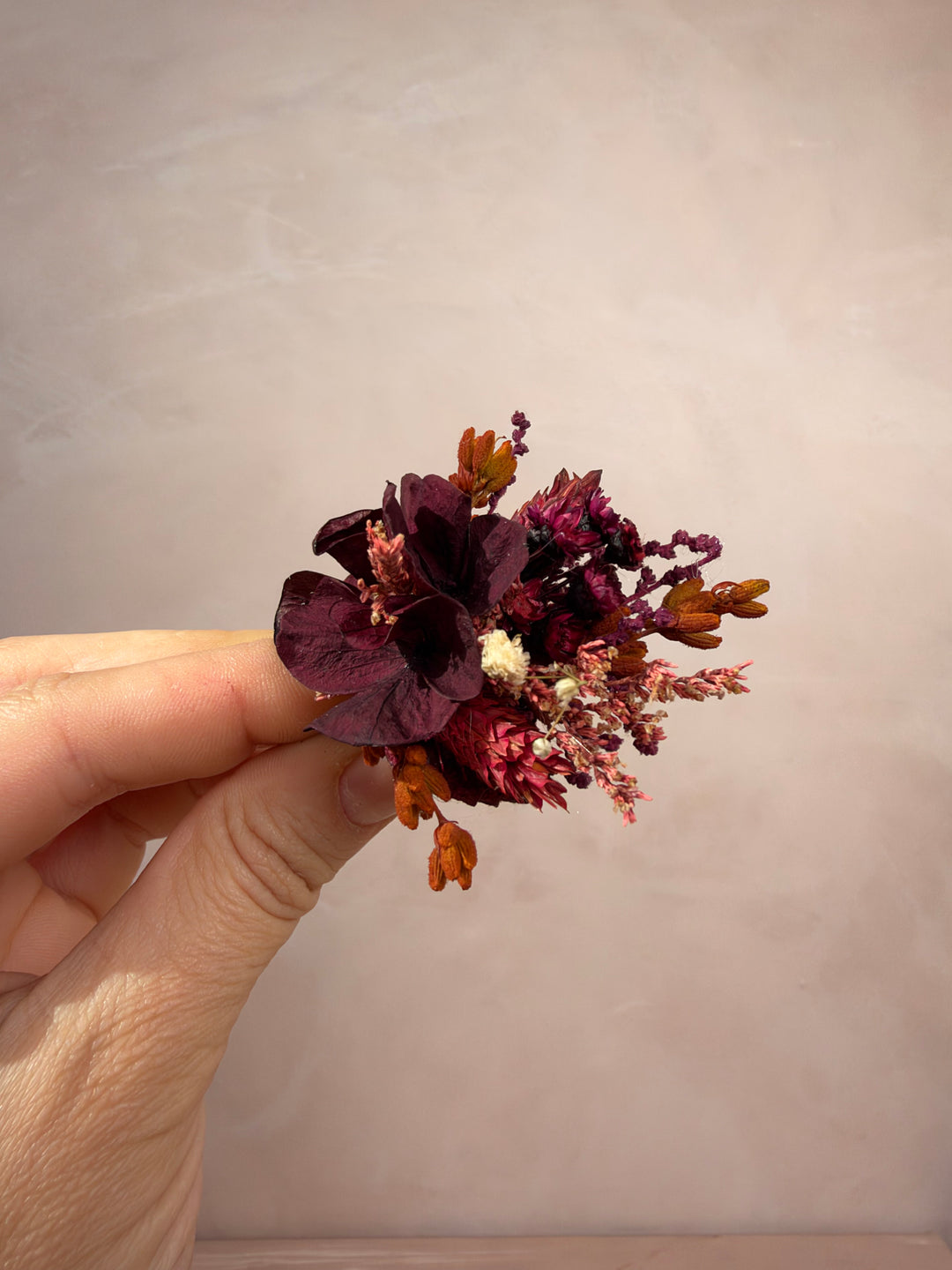 A hand holding a small dried floral ring featuring deep burgundy petals, orange-brown buds, and tiny cream accents against a soft beige background.