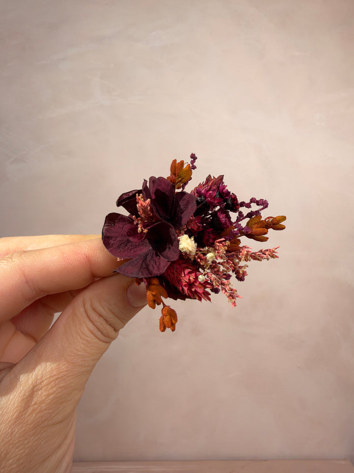 A hand holding a small dried floral ring featuring deep burgundy petals, orange-brown buds, and tiny cream accents against a soft beige background.