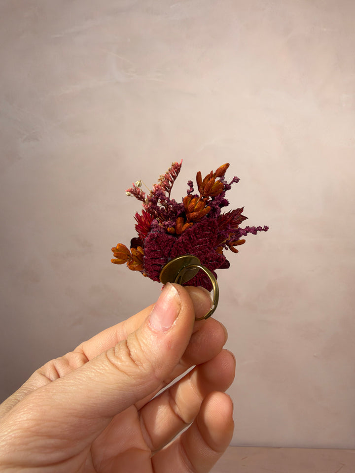 A hand holding a small dried floral ring with burgundy and orange flowers set in a gold-toned band, against a soft beige background.