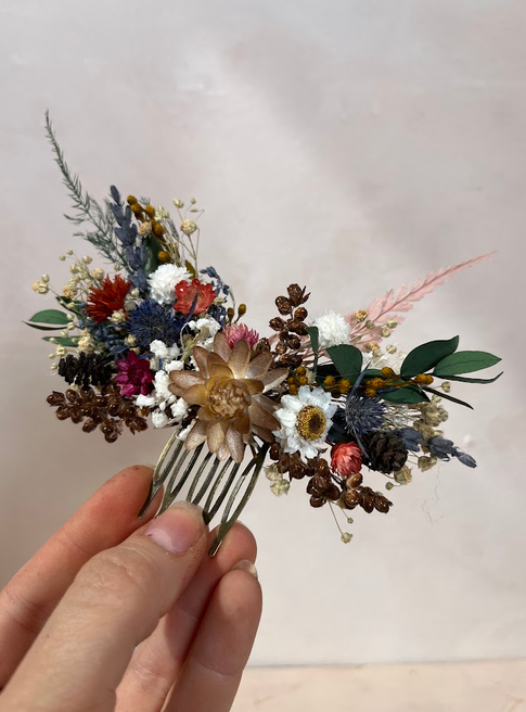 A hand holds a dried floral hair comb featuring a variety of small flowers, green leaves, and pinecones against a soft, light beige background.