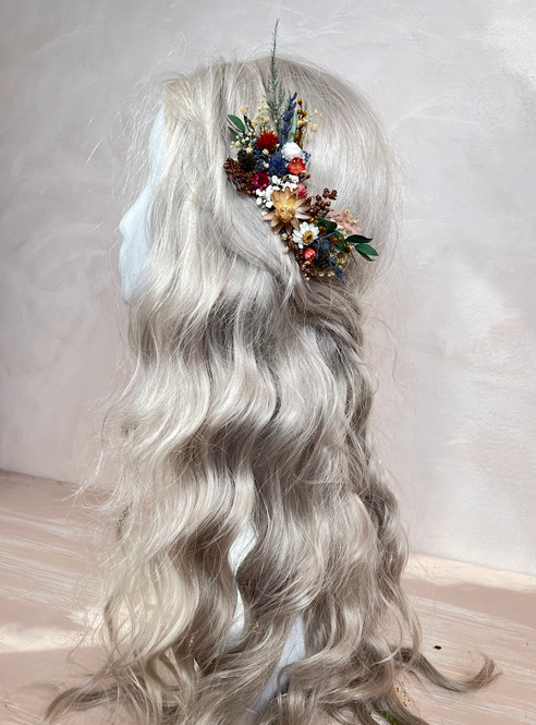 Long wavy platinum blonde wig with a small multicolor dried flower hair accessory, displayed on a mannequin head against a neutral light beige background.