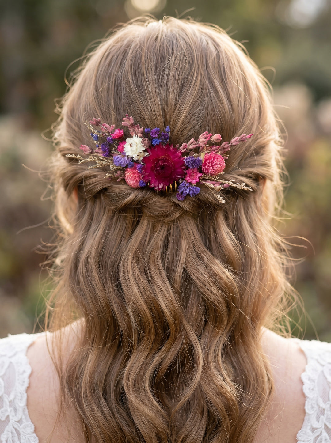 A woman viewed from behind with long wavy light brown hair, wearing a floral hair accessory with pink, purple, and white flowers in a half-up bun.