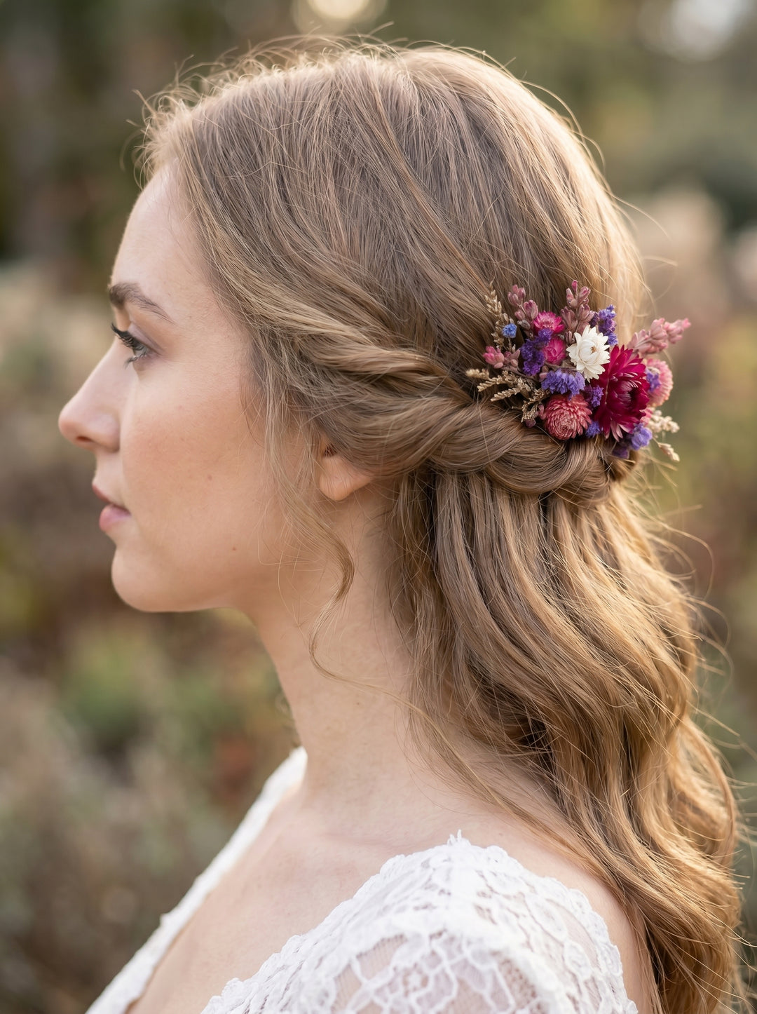 A woman in profile wearing a delicate floral hair accessory with pink, purple, and white dried flowers in her half-updo hairstyle, set against a soft, blurred outdoor background.