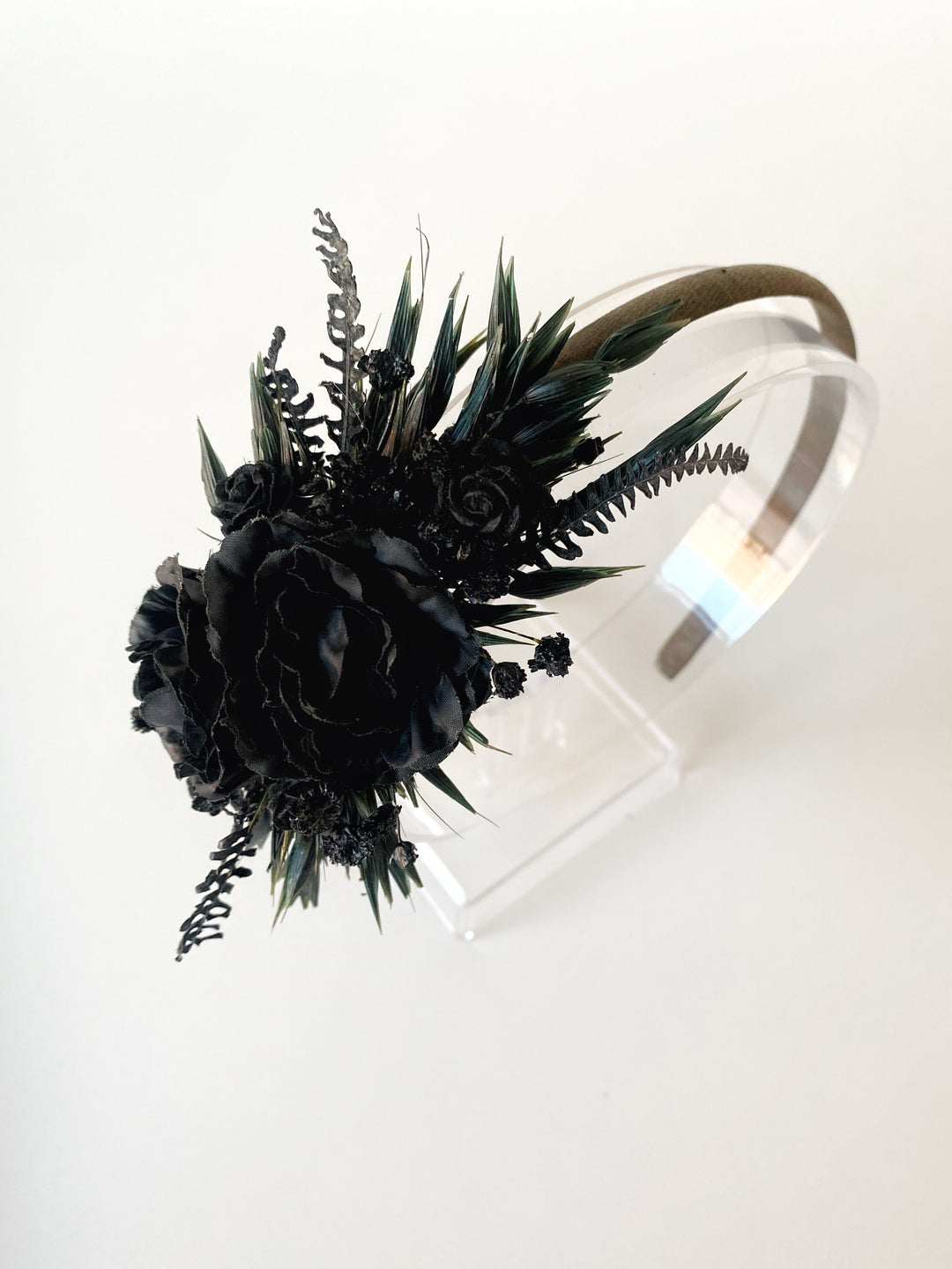 A black floral headband with a large black rose and dark green leaves, displayed on a clear acrylic stand against a white background.