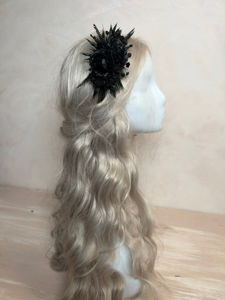 A long wavy platinum blonde wig displayed on a white mannequin head, styled with a black feather and floral headpiece, set against a neutral beige background.