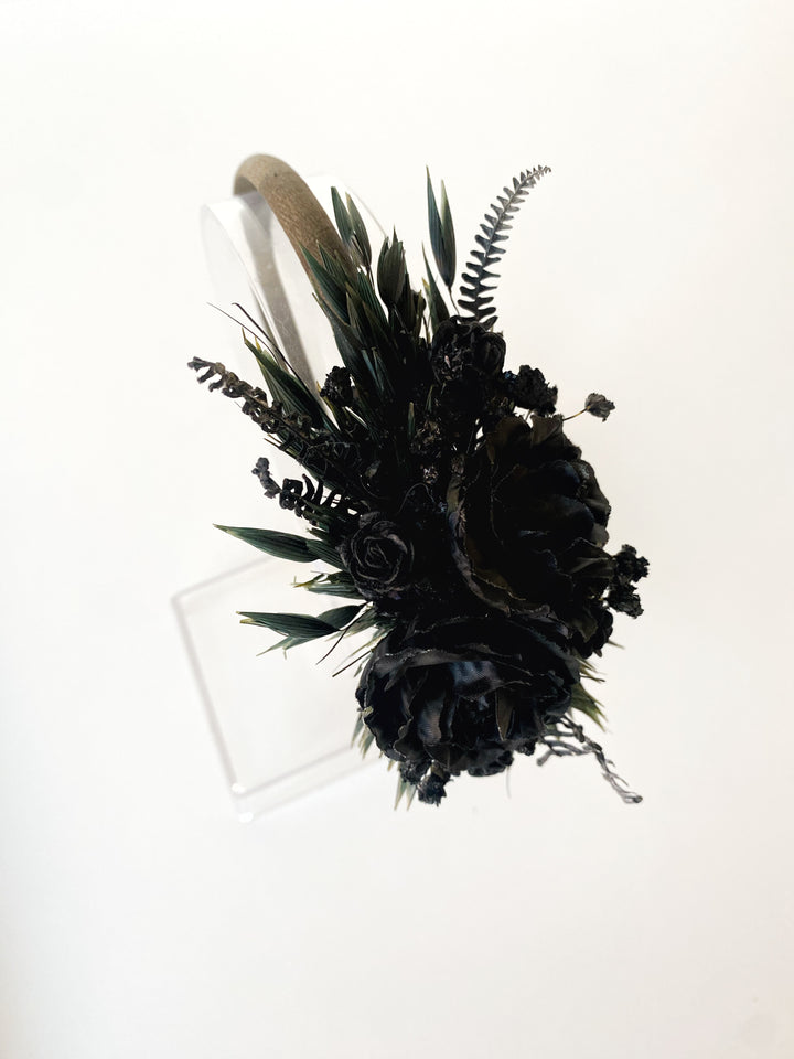A black floral  headband featuring black roses, ferns, and green leaves, attached to a beige fabric wristband, displayed on a clear acrylic stand against a white background.
