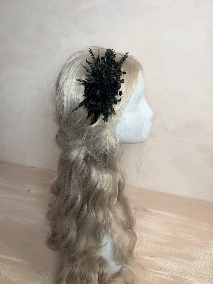 A long wavy blonde wig displayed on a white mannequin head, styled with a black feathered floral hair accessory, set against a neutral beige background.