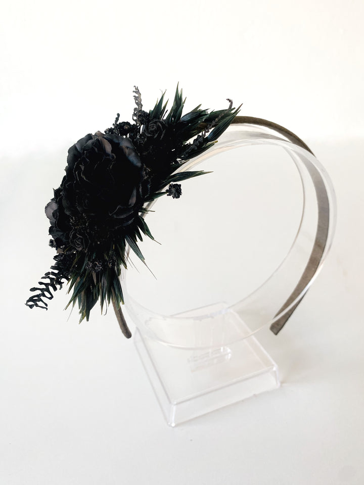 A black floral headband with dark flowers, leaves, displayed on a clear acrylic stand against a white background.