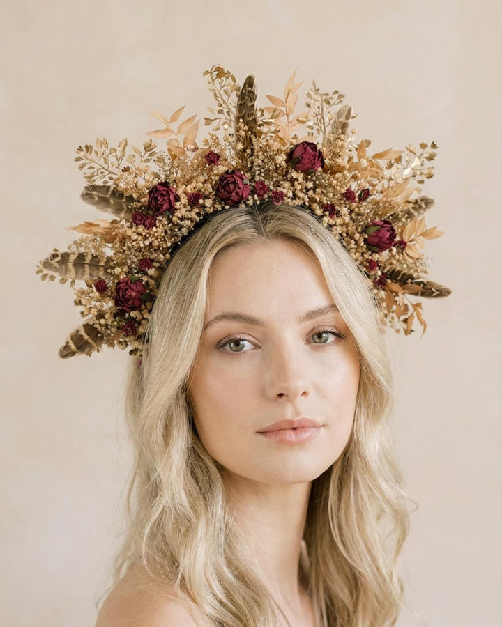 Woman wearing a floral crown with burgundy flowers, feathers and gold leaves on a plain background