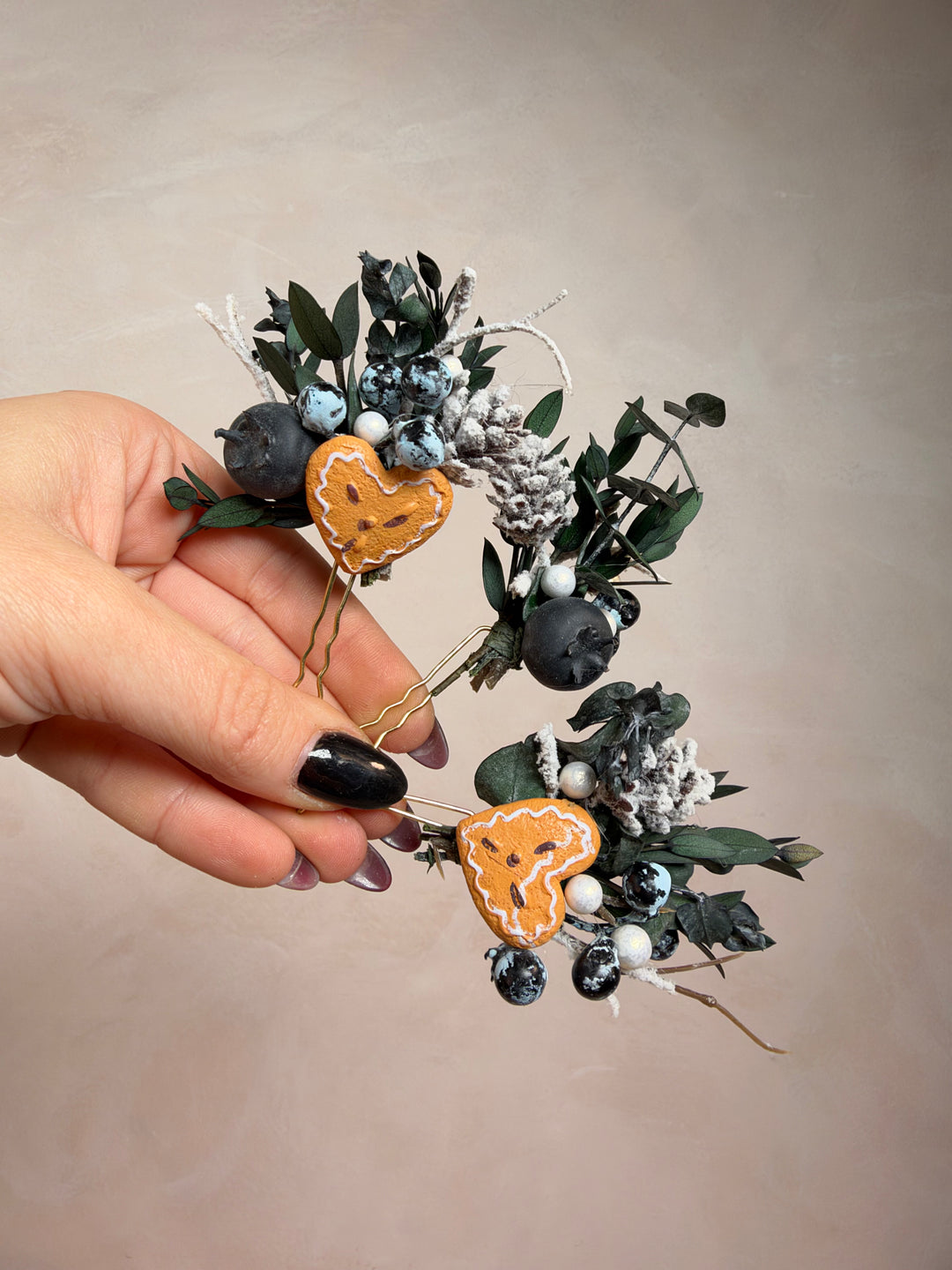 A hand with black nail polish holds a dried floral arrangement featuring dark green eucalyptus leaves, faux blueberries, frosted white berries, and two small orange heart-shaped accents against a plain beige background.
