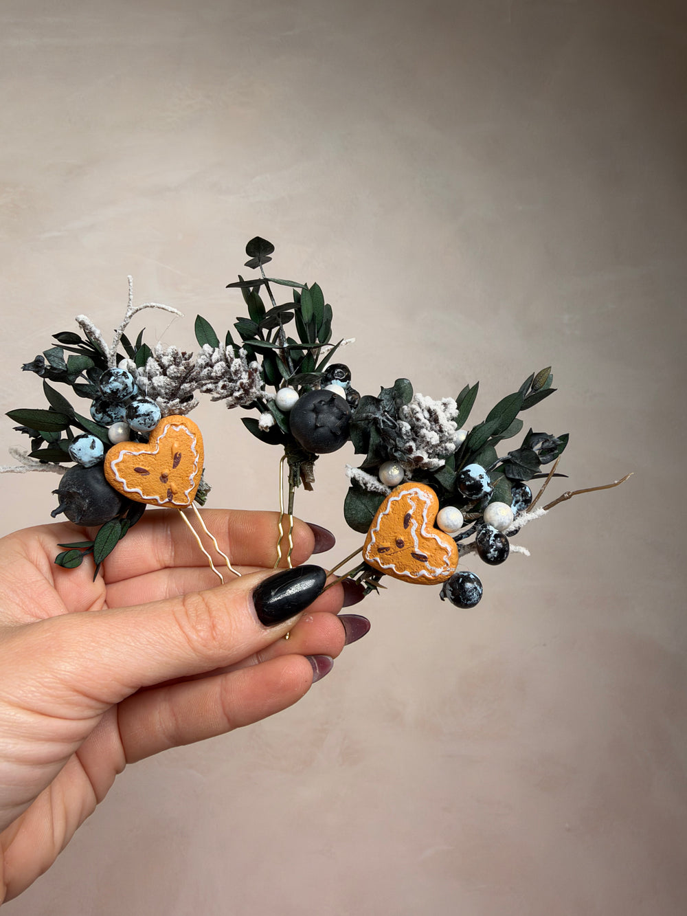 A hand with black nail polish holds a small decorative floral arrangement featuring two gingebread hearts, dark green leaves, white berries, and dark blue berries against a plain beige background.