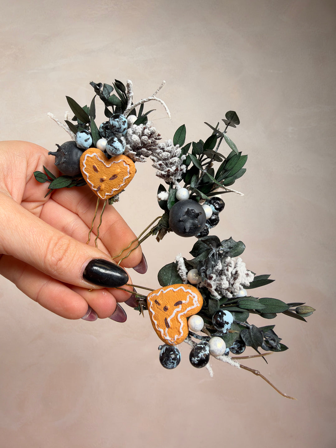 A hand with black nail polish holds a small decorative floral arrangement featuring two faux gingebread hearts, dark blue berries, white berries, green leaves, and frosted pinecones against a plain beige background.
