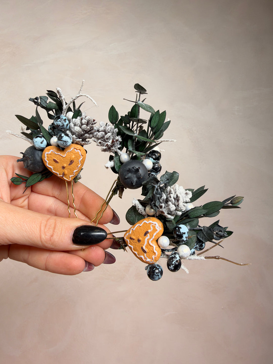 A hand with black nail polish holds a decorative wreath made of green eucalyptus leaves, white faux snow, dark berries, and two orange pumpkin-shaped ornaments, against a plain beige background.