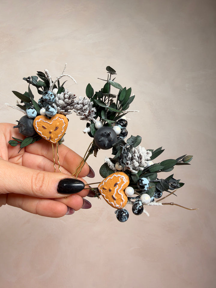 A hand with black nail polish holds a decorative wreath made of green eucalyptus leaves, white faux snow, dark berries, and two orange pumpkin-shaped ornaments, against a plain beige background.