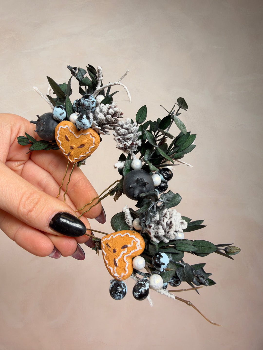 A hand with black nail polish holds a decorative dried floral arrangement featuring green eucalyptus leaves, white berries, dark gray stones, and two gingebread hearts against a plain beige background.