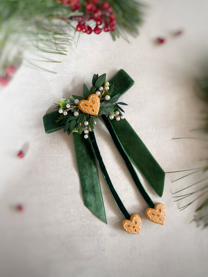 A dark green velvet hair bow accessory with a decorative floral cluster featuring green leaves, white berries, and gingerbread heart-shaped charms, displayed on a light neutral background with festive pine and red berries.