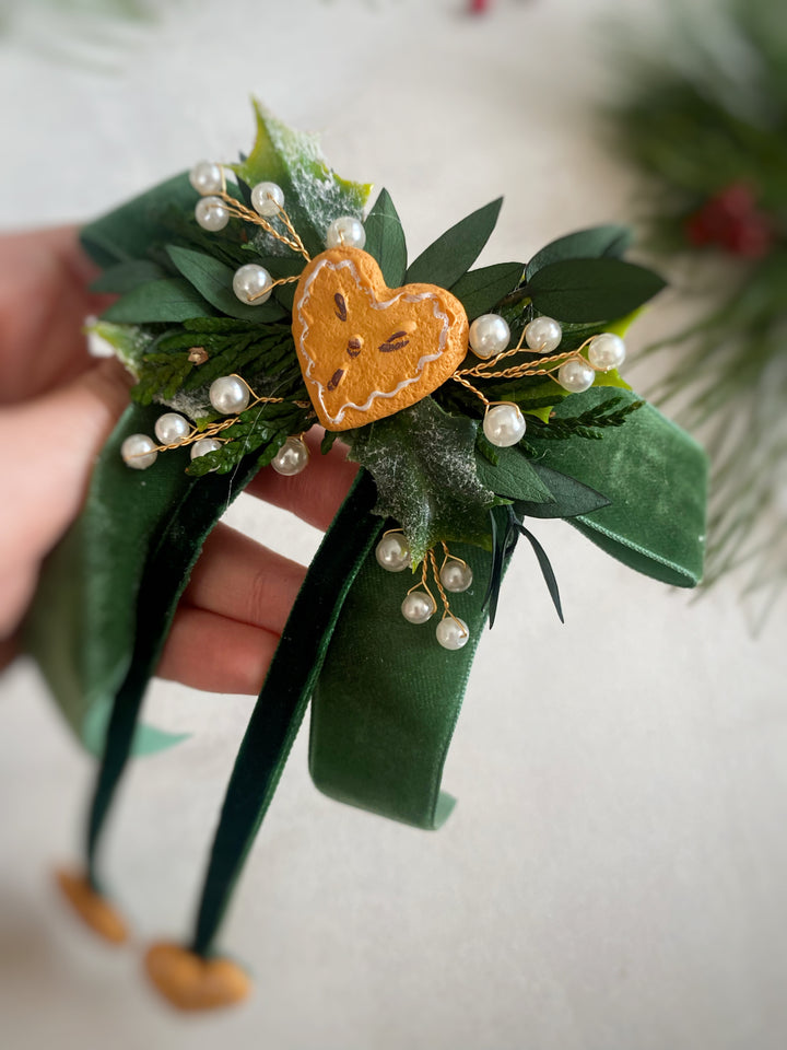 A hand holds a green velvet headband decorated with artificial and preserved green leaves, small white pearls, and a central orange-brown felt heart gingerbread ornament. 