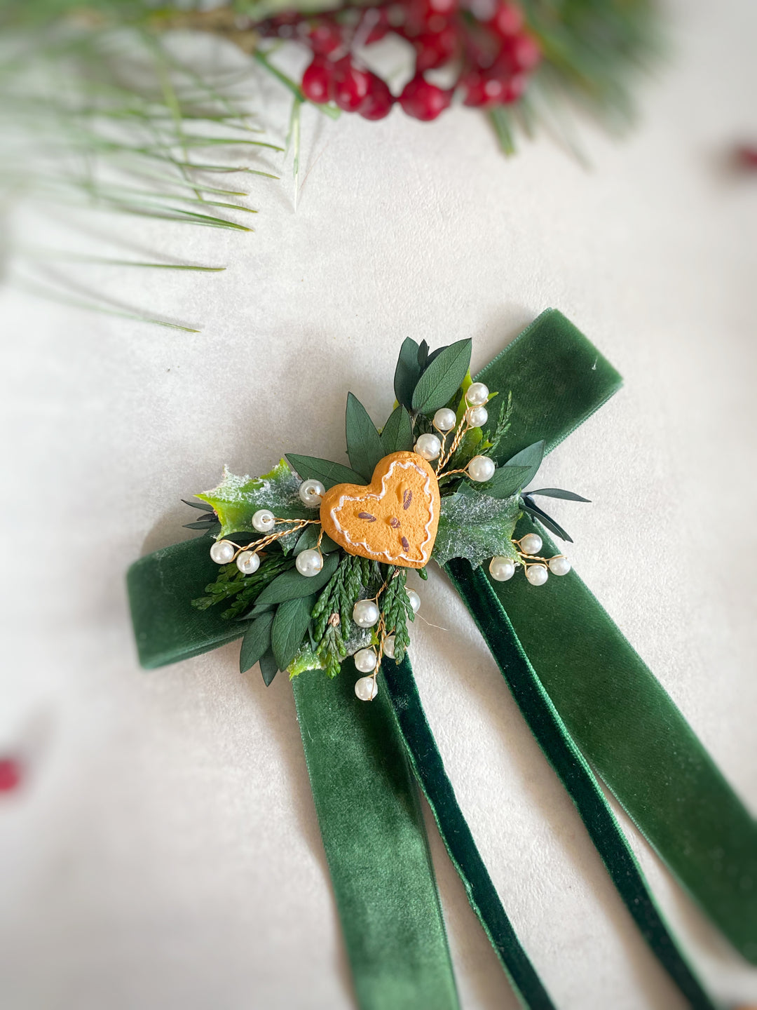 A green velvet ribbon corsage with a small holly leaf, white pearl beads, and a central gingebread heart, displayed on a light textured background with pine branches and red berries.