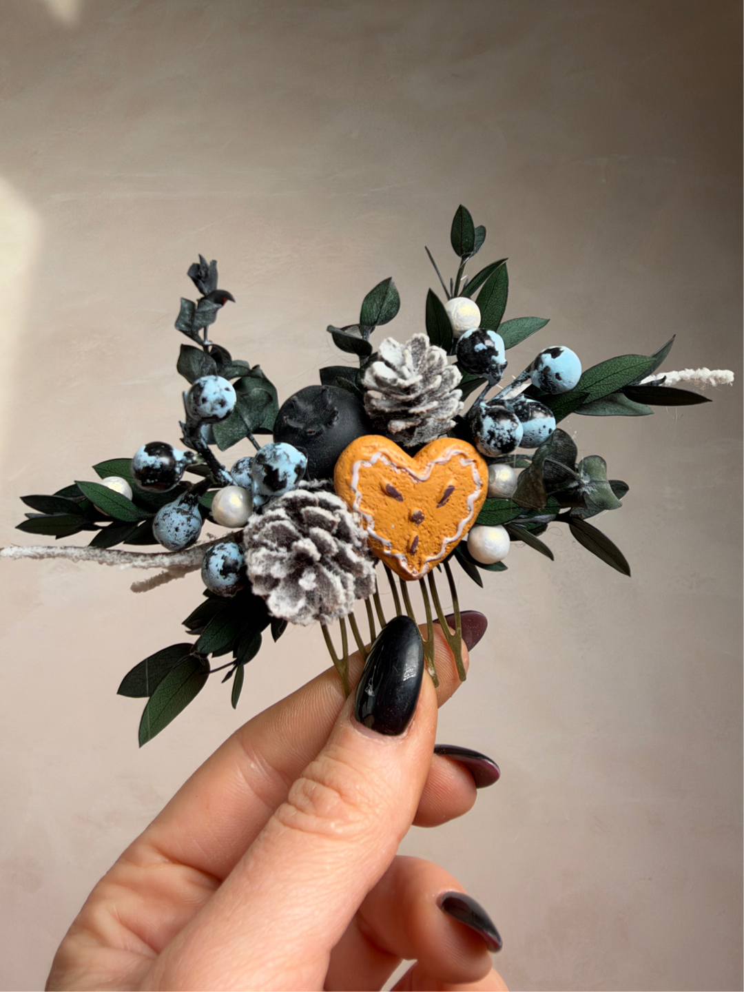 A hand with black nail polish holds a decorative hair comb featuring a small heart, white and blue berries, green leaves, and faux pinecones against a plain beige wall.
