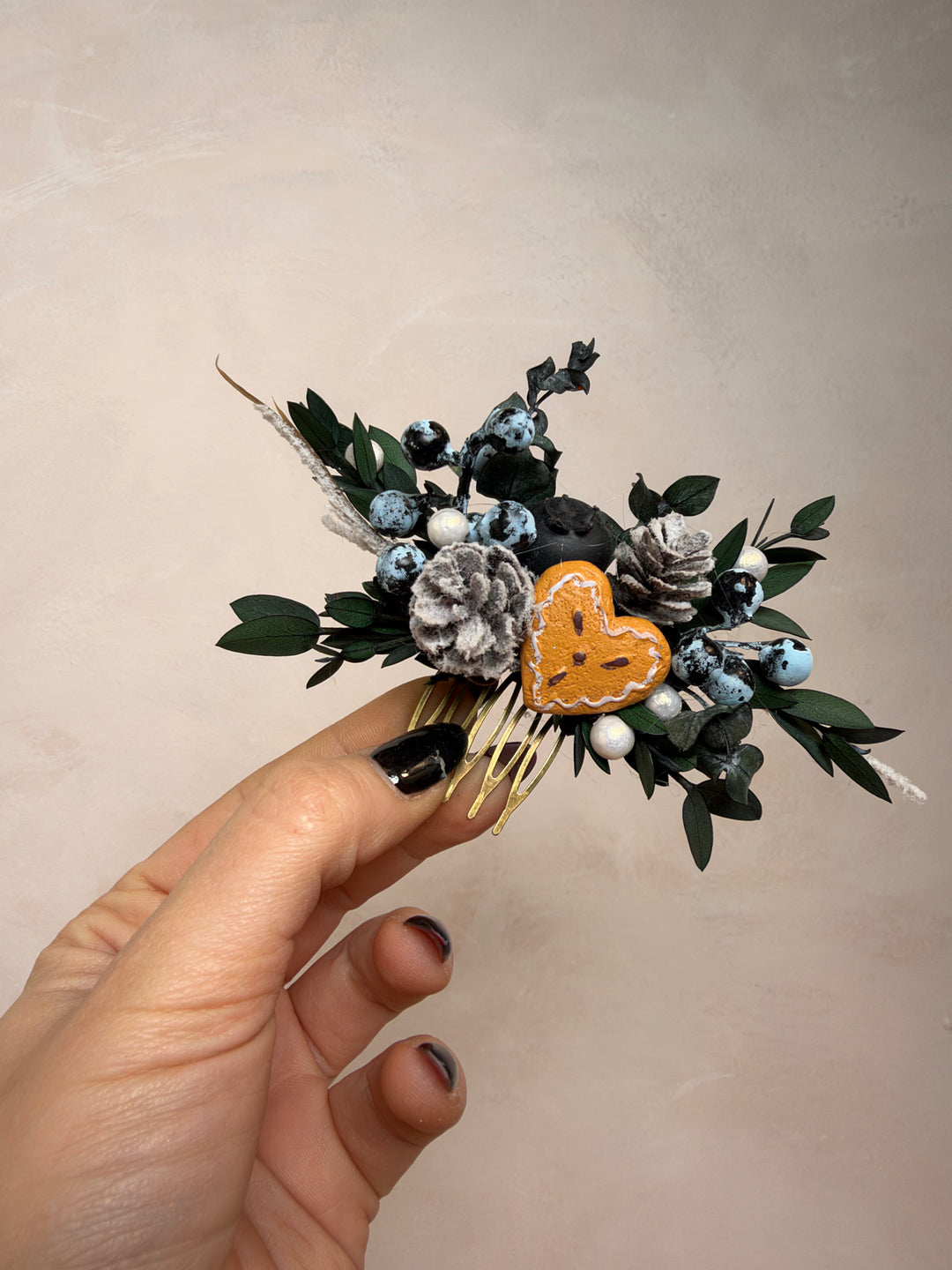 A hand with black nail polish holds a small decorative hair accessory featuring a gingerbread heart, pinecones, blue berries, white beads, and green leaves on a gold comb, set against a neutral beige background.