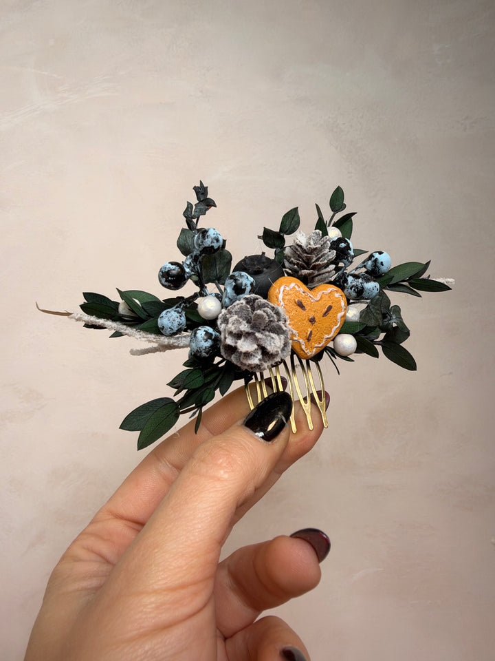 A hand with black nail polish holds a small decorative hair comb featuring dark green leaves, blue berries, a gray textured flower, and a central heart-shaped accent on a plain beige background.