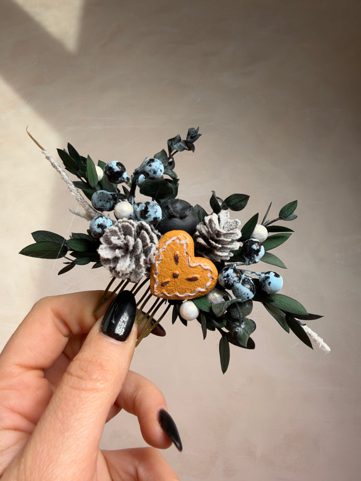 A hand with black nail polish holds a small decorative hair accessory featuring a central heart, white and blue berries, green leaves, and pinecone-like floral elements against a plain beige wall.