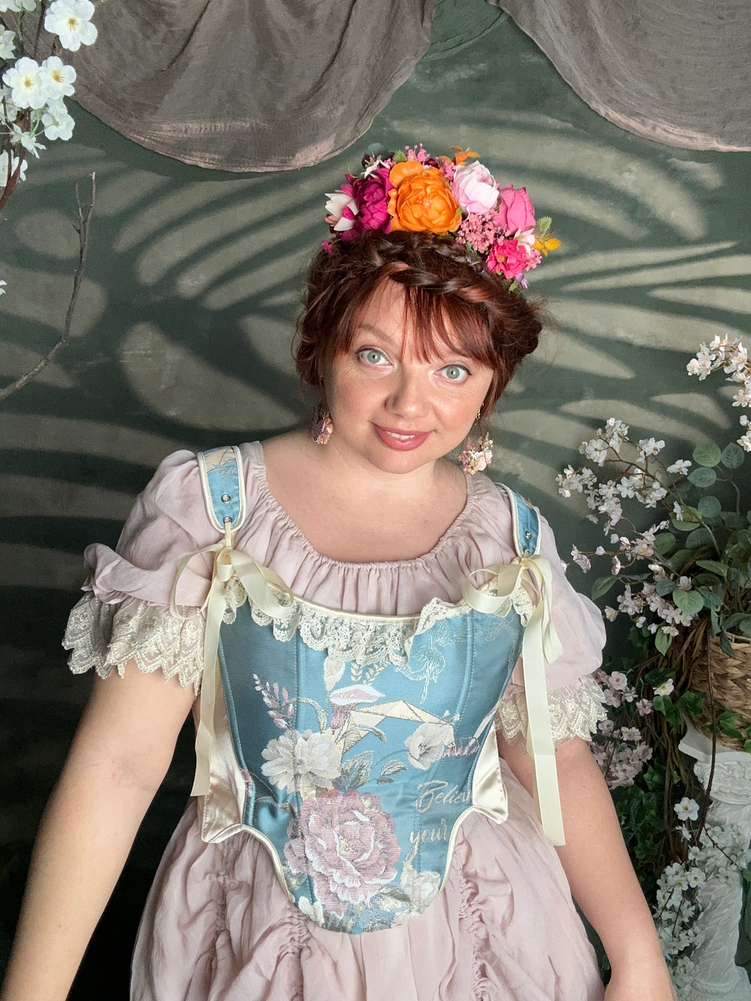 A woman wearing a vintage-style light blue and pink floral dress with lace trim and a matching flower crown, photographed in a decorative garden-like setting.