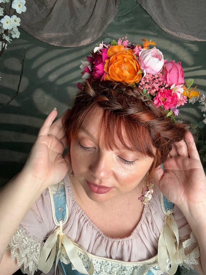 A woman with closed eyes wearing a colorful floral crown of pink, orange, and white flowers, dressed in a light pink ruffled top, lying down against a dark gray background.