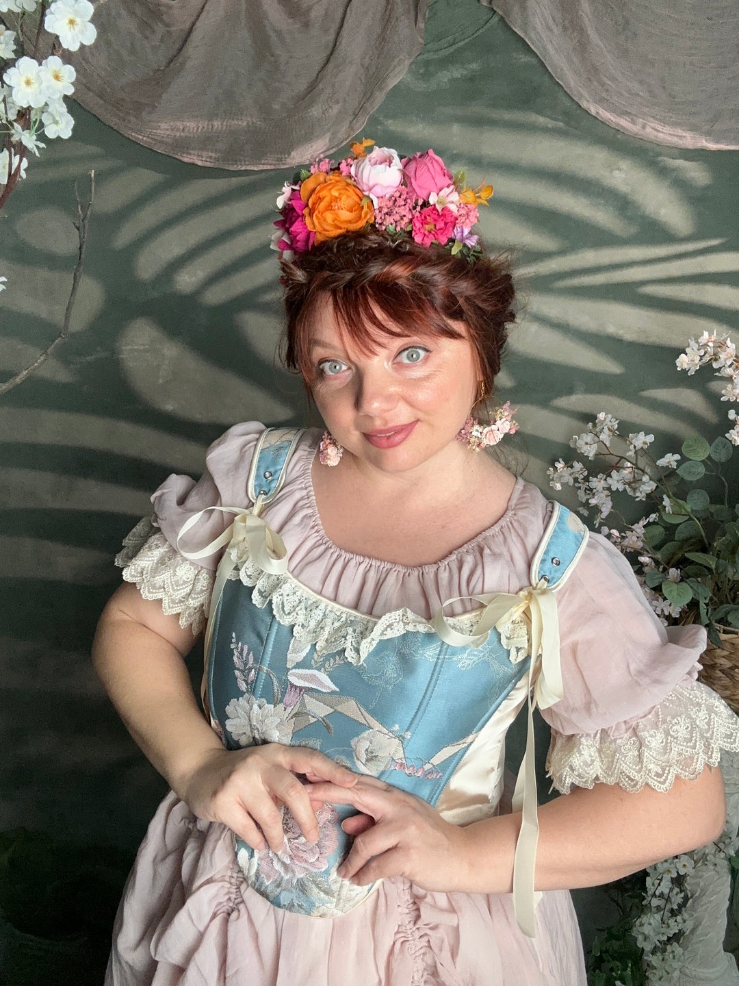 A woman wearing a vintage-style pastel dress with lace trim, a light blue apron-like bodice panel, and a pink skirt, posed against a dark green backdrop with floral decorations.
