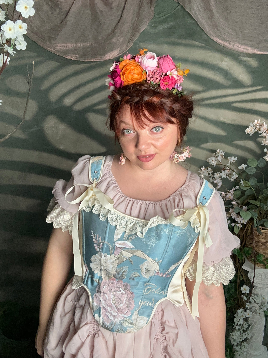 A woman wearing a vintage-style lavender dress with a light blue floral apron overlay and a pink and orange flower crown, photographed in a garden-like setting with cast shadows.