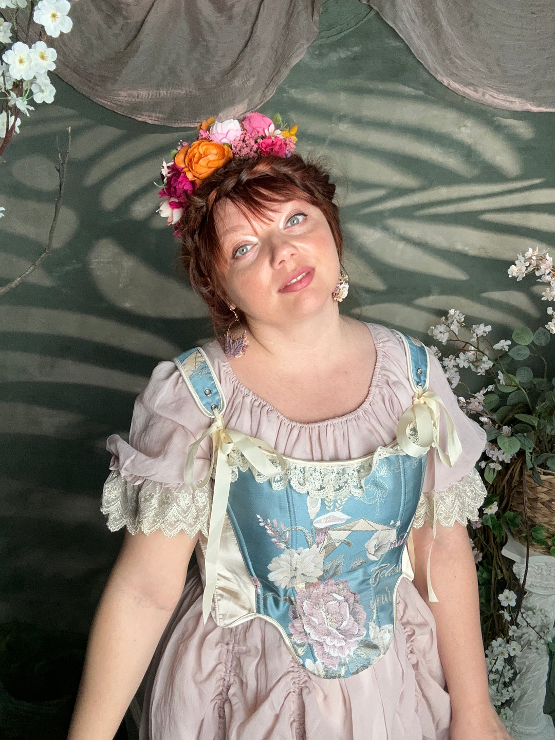 A woman wearing a light pink and blue floral Victorian-style dress with lace trim, a matching flower crown, and earrings, photographed in a garden setting.