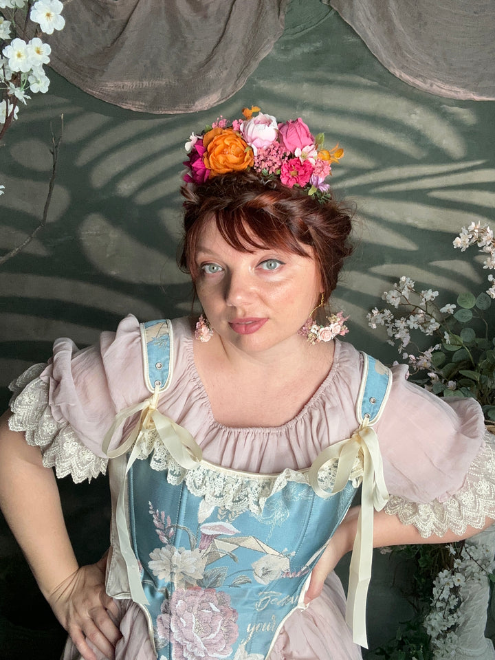 A woman wearing a vintage-style pastel dress with a floral headpiece, photographed against a dark green backdrop with leaf shadows and white flowers.