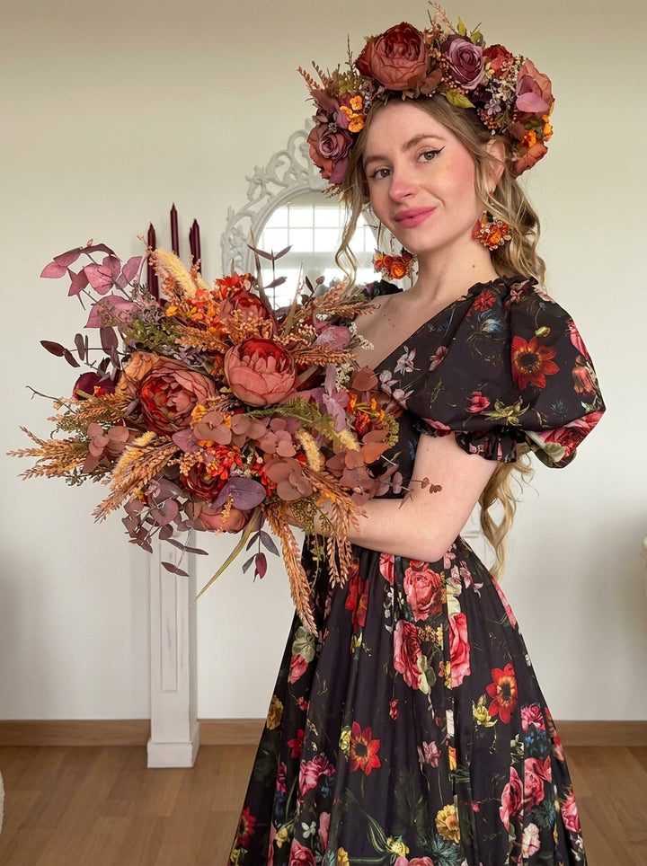 A woman wearing a black floral dress and a large flower crown holds a large autumn floral bouquet in a bright indoor room.