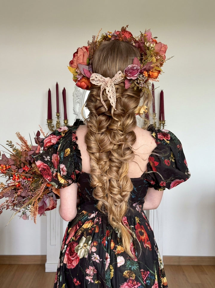 A woman viewed from behind wearing a long, elaborate braided hairstyle with a large floral crown and bouquet, in a black floral dress against an indoor background with candles.
