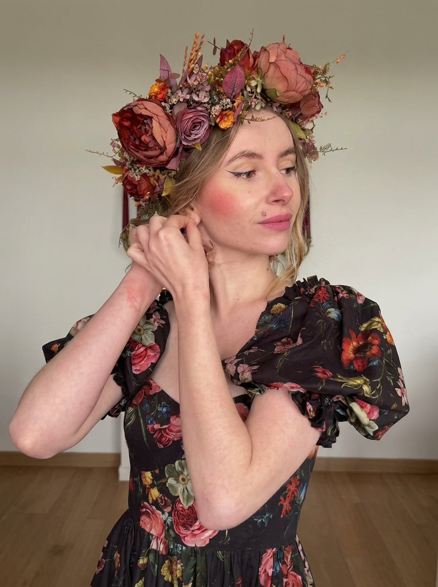 A woman wearing a large, rustic flower crown with muted red, pink, and orange blossoms, paired with a black floral puff-sleeve dress, posing indoors against a plain wall.