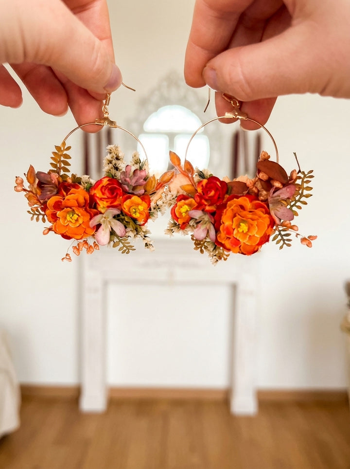A pair of large gold hoop earrings adorned with orange, red, and yellow flowers and green leaves, held up by two hands against a softly blurred indoor background.