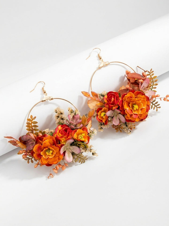 A pair of large gold-tone hoop earrings decorated with orange, red, and pink flowers and green-brown leaves, displayed on a clean white background.