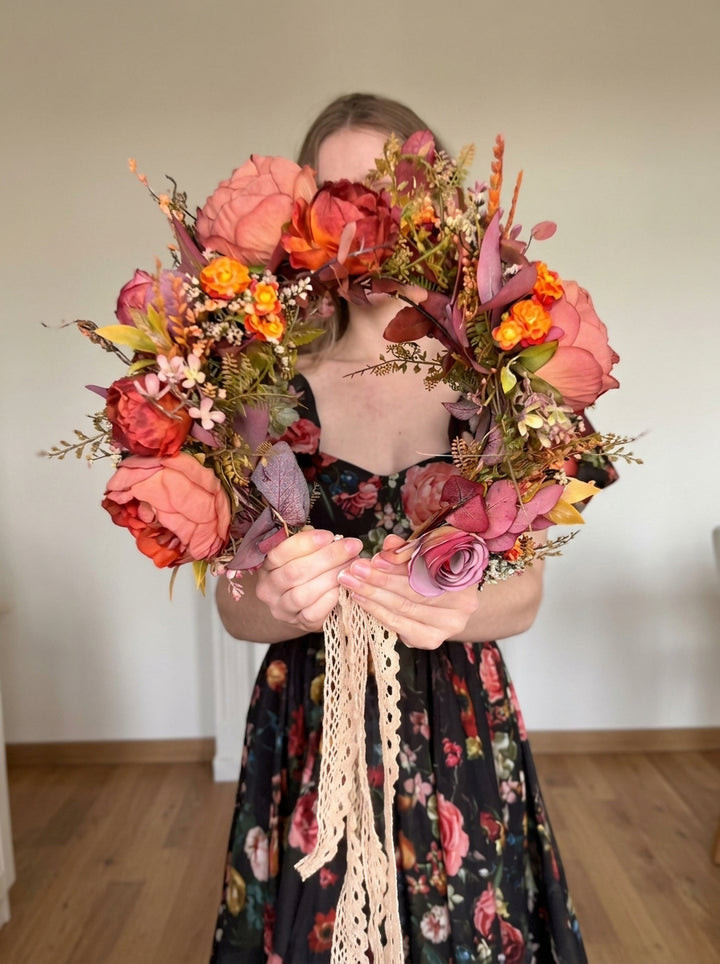 A woman in a floral dress holds a large circular floral wreath made of pink, orange, and burgundy faux flowers with greenery and a cream lace ribbon, photographed indoors against a plain wall and wood floor.