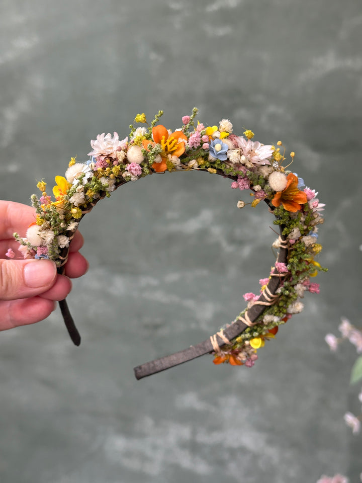 A handmade floral headband decorated with orange, yellow, pink, and white flowers, held by a hand against a textured gray background.