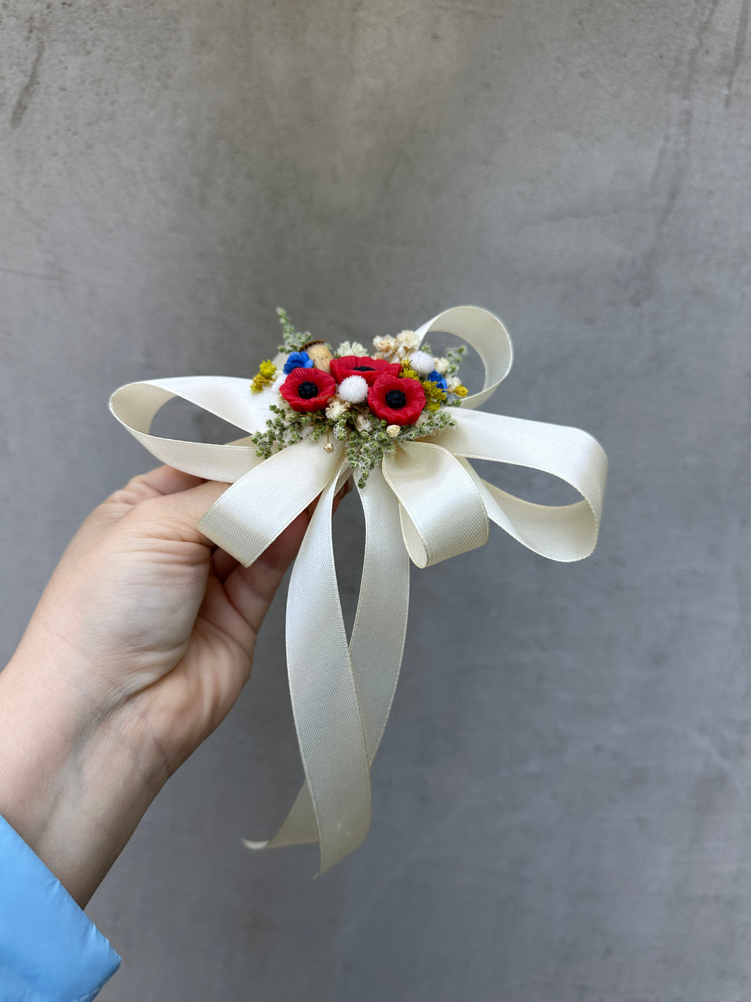 A hand holding a small decorative flower arrangement with a large cream ribbon bow. The center features red poppy-like flowers, small white blossoms, and green foliage against a textured gray wall.