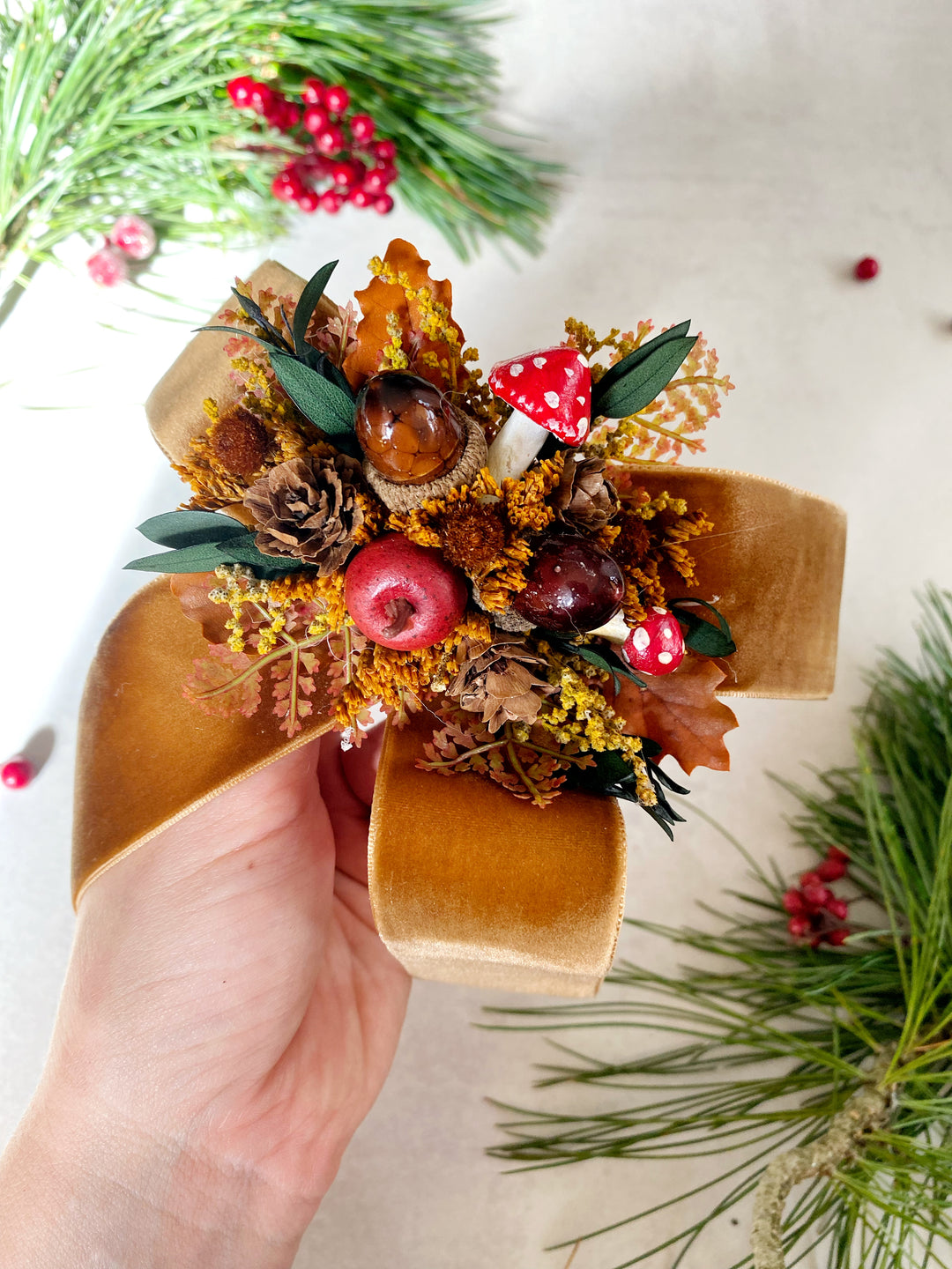 A hand holds a decorative hair bow  featuring autumn leaves, acorns, pinecones, small red berries, and two tiny red-and-white polka dot mushrooms, styled with pine branches on a light neutral background.