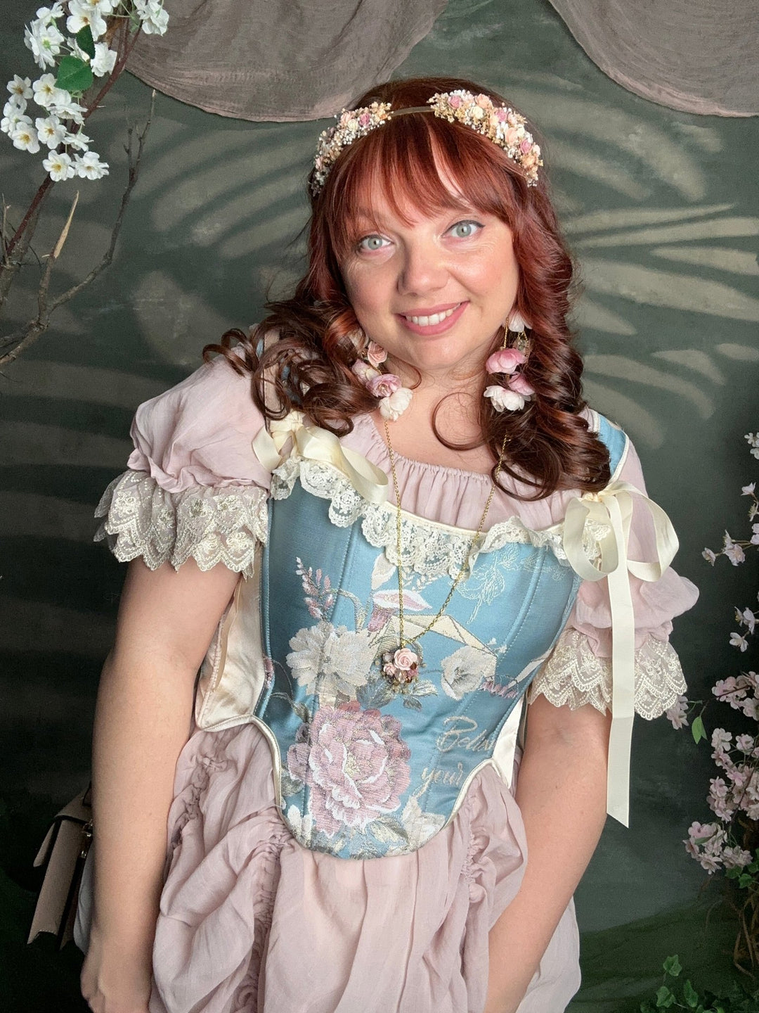 A woman wearing a vintage-style medieval or Victorian costume dress with a light blue floral corset bodice, pale pink ruffled sleeves, and a matching pink skirt, posed against a dark green backdrop with floral decorations.