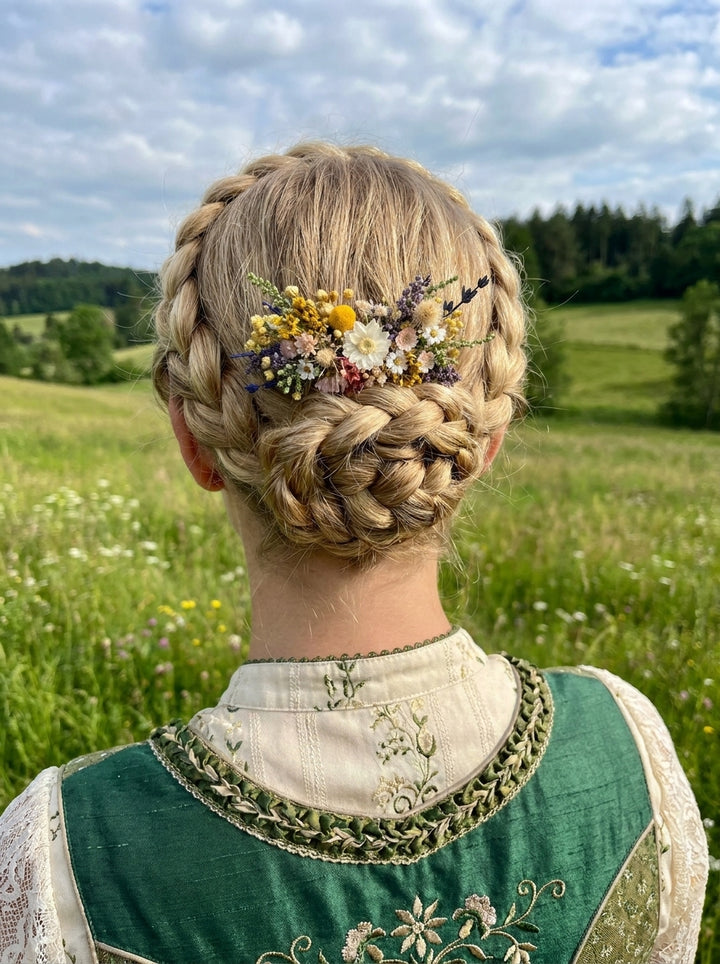 A blonde woman viewed from behind outdoors in a grassy field, wearing a green embroidered traditional-style top and a braided hairstyle adorned with a small multicolor floral hair accessory.