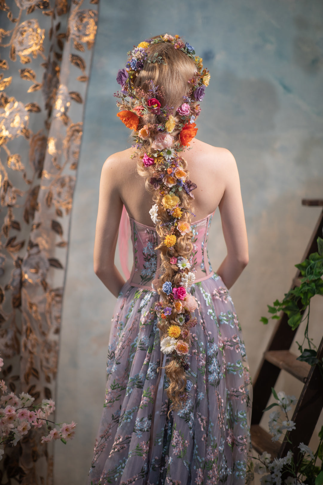 Woman in a floral dress with a long floral hairpiece against a textured wall.