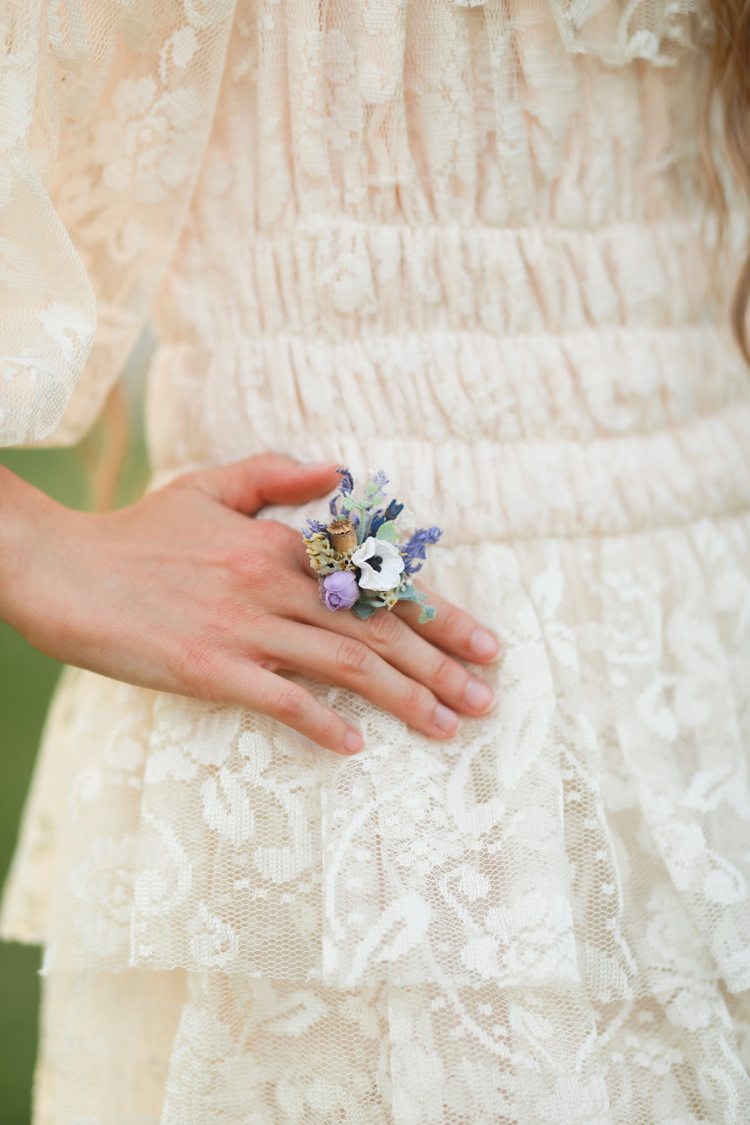 Close-up of a hand holding a small floral purple ring against a lace dress background